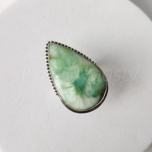 May include: A silver ring with a green gemstone in a teardrop shape. The gemstone has a white and green pattern.