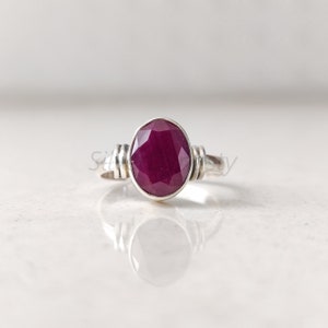 May include: A silver ring featuring an oval-cut, deep red ruby gemstone. The ring has a simple band with decorative silver accents on either side of the ruby. The gemstone is the focal point, set in a polished silver bezel.