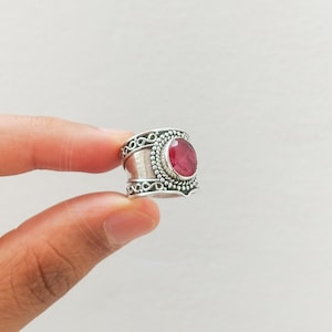 May include: A silver ring featuring a red gemstone set in an ornate bezel. The ring has a wide band with intricate detailing.