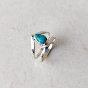 May include: A silver ring with a turquoise teardrop-shaped stone set in the center. The ring is designed with two bands that wrap around the finger.