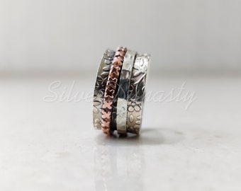 May include: A wide spinner ring featuring a central copper band with a repeating cross pattern, flanked by two silver bands. The outer silver bands are decorated with an embossed floral motif, while the inner silver band has a hammered texture.