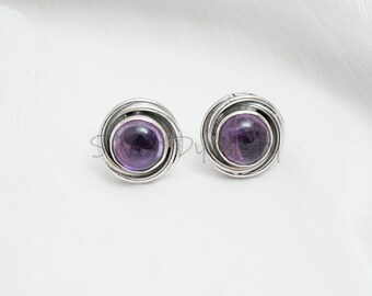 May include: A pair of silver stud earrings with a purple amethyst gemstone in each. The earrings have a simple, elegant design with a circular setting.