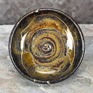 May include: A round ceramic bowl featuring a glossy glaze with a swirling pattern of deep black, dark brown, and golden yellow tones. The interior surface displays a prominent spiral design radiating from the center, with a smooth, reflective finish.