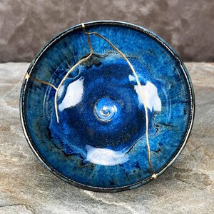 May include: A circular ceramic bowl featuring a deep blue glaze with lighter blue and black variations. The bowl displays a prominent crack repaired with a gold-colored material, creating a distinct line that extends across the interior surface.