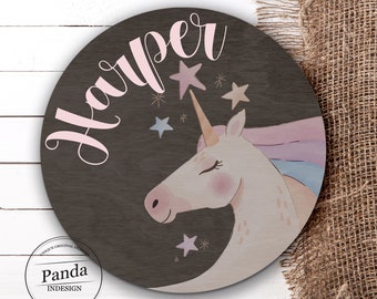 May include: A round wooden sign with a dark brown background featuring a white unicorn with a pink horn and mane. The unicorn is surrounded by pink, blue, and white stars. The name "Harper" is written in pink cursive letters at the top of the sign. The sign also includes the text "Panda InDesign" in a white circle with a black border.