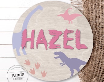 Może przedstawiać: Okrągły drewniany znak z motywem różowych dinozaurów. Znak zawiera imię "HAZEL" w różowych literach z efektem postarzenia. Znak zawiera również sylwetki fioletowych dinozaurów, różowe ślady dinozaurów i różowo-białe logo z napisem "Panda Indesign".