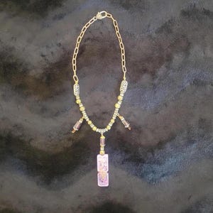 May include: A gold-colored chain necklace with a pendant. The necklace has a series of small, round beads and rectangular, pinkish-purple stones. The pendant is a vertical rectangle with two stacked, smaller rectangles. The background is a dark, textured surface.