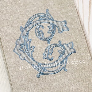 May include: A linen tea towel with a blue embroidered letter G in a decorative floral design. The text "Alabama Honey Designs" is visible in the bottom right corner. The text "Smokey blue on linen" is visible in the bottom left corner.