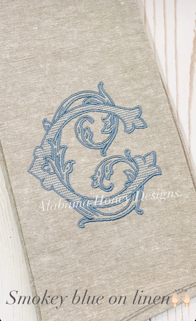 May include: A linen tea towel with a blue embroidered letter G in a decorative floral design. The text "Alabama Honey Designs" is visible in the bottom right corner. The text "Smokey blue on linen" is visible in the bottom left corner.