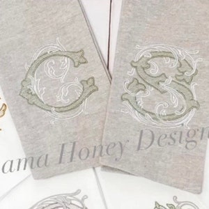 May include: Two embroidered towels with a decorative monogram design. The towels are a light grey colour and the monogram is in a light green and white thread. The text on the towel reads "Alabama Honey Designs".