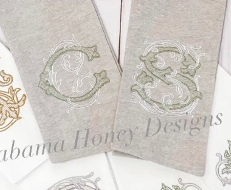 May include: Two embroidered towels with a decorative monogram design. The towels are a light grey colour and the monogram is in a light green and white thread. The text on the towel reads "Alabama Honey Designs".