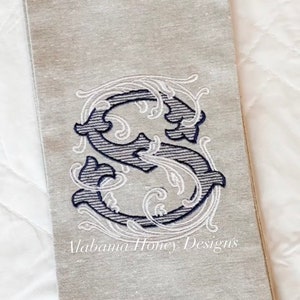 May include: A linen tea towel featuring a navy and white embroidered letter "S" with decorative flourishes. The text "Alabama Honey Designs" is embroidered below the letter. The towel is on a white surface.