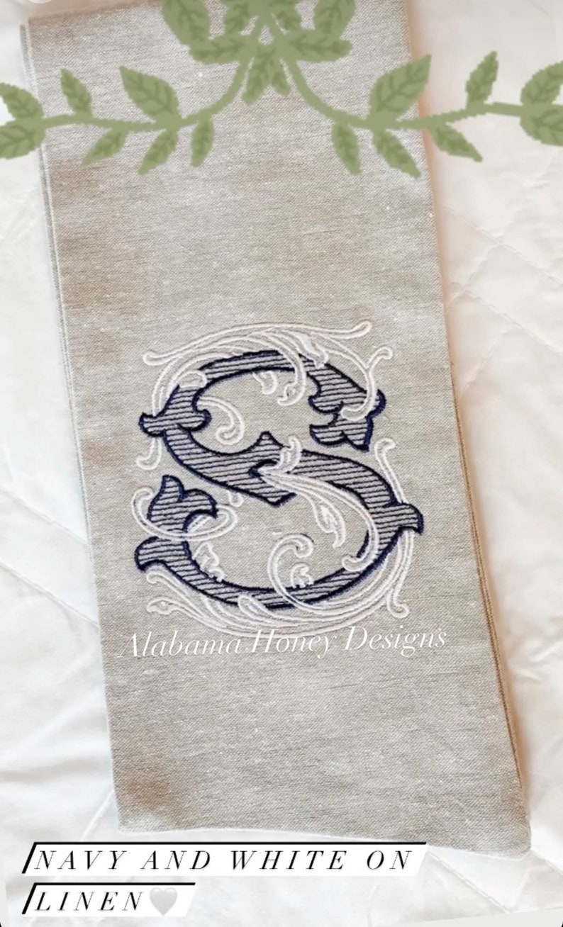 May include: A linen tea towel featuring a navy and white embroidered letter "S" with decorative flourishes. The text "Alabama Honey Designs" is embroidered below the letter. The towel is on a white surface.