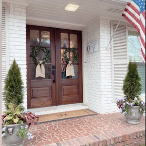 May include: A white brick house with a brown double door entrance. The door has a wreath on each side and a welcome mat. There are two potted plants on either side of the door. An American flag is flying on a pole to the right of the house.