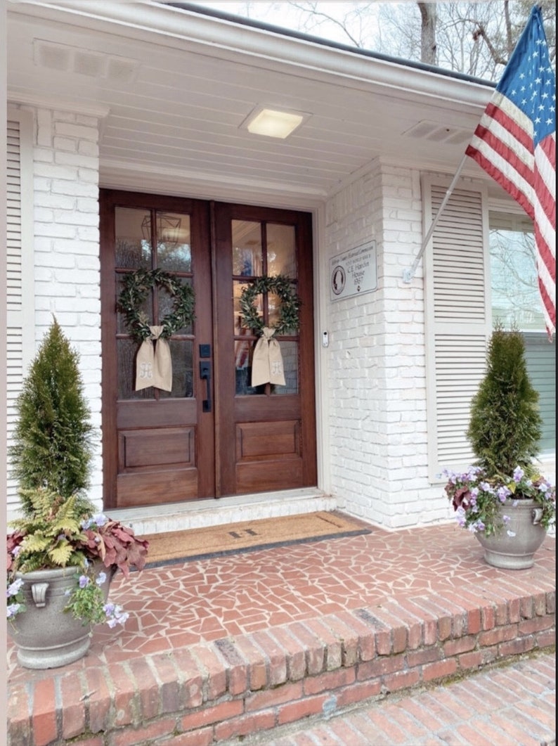 May include: A white brick house with a brown double door entrance. The door has a wreath on each side and a welcome mat. There are two potted plants on either side of the door. An American flag is flying on a pole to the right of the house.