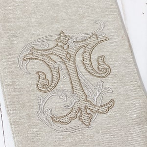 May include: A beige linen tea towel with a brown embroidered monogram of the letter "T" in a decorative script font.