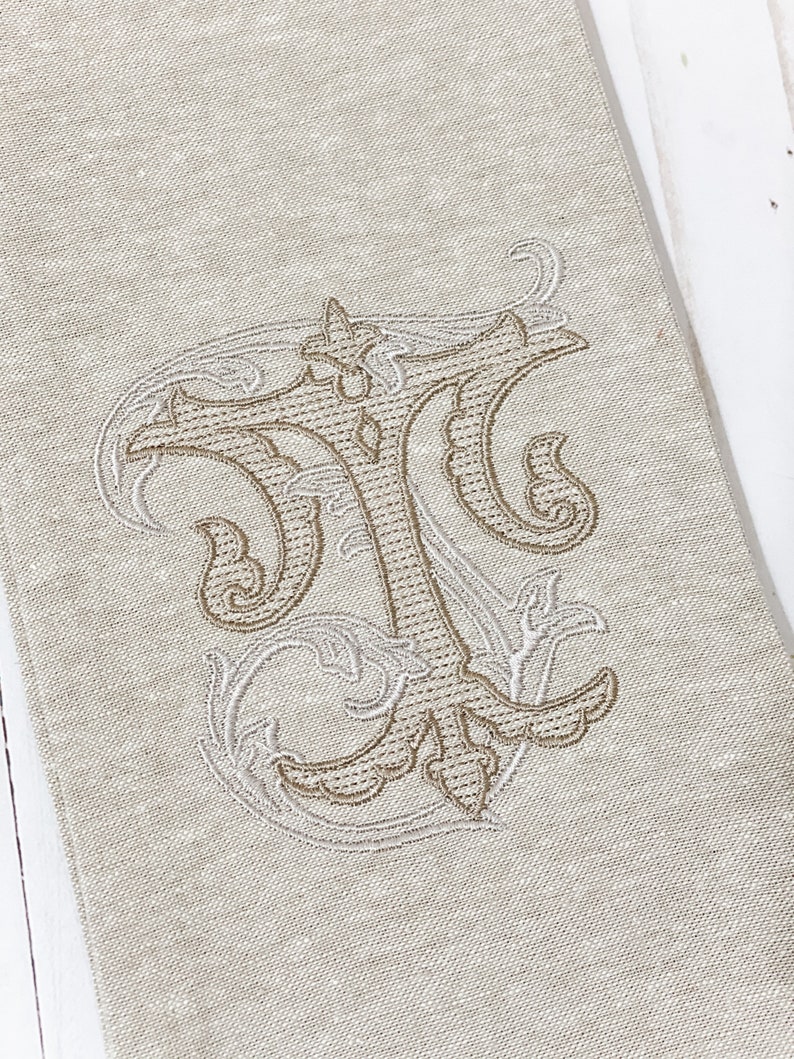 May include: A beige linen tea towel with a brown embroidered monogram of the letter "T" in a decorative script font.