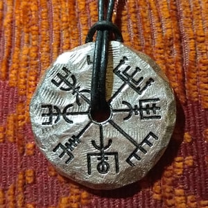 May include: A silver pendant with a compass design featuring Norse runes. The pendant is attached to a black leather cord.