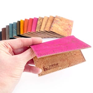 Pu&ograve; includere: Un porta carte in sughero rosa e naturale &egrave; tenuto aperto, mostrando l'interno. Una variet&agrave; di portafogli in sughero colorati sono a ventaglio sullo sfondo. Il porta carte ha il logo "minter GOODS".