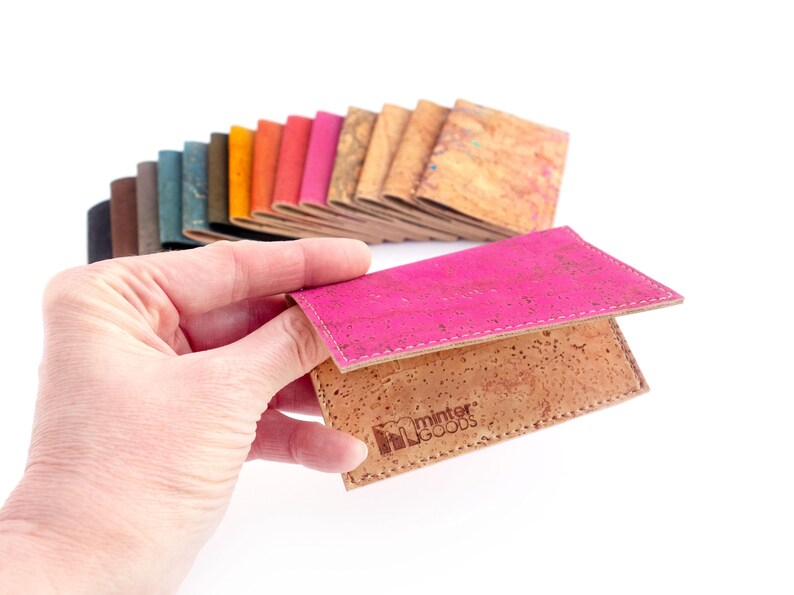 Pu&ograve; includere: Un porta carte in sughero rosa e naturale &egrave; tenuto aperto, mostrando l'interno. Una variet&agrave; di portafogli in sughero colorati sono a ventaglio sullo sfondo. Il porta carte ha il logo "minter GOODS".