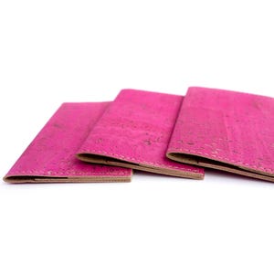Pu&ograve; includere: Tre portafogli in sughero rosa acceso sono esposti su sfondo bianco. I portafogli sono aperti, rivelando un interno marrone chiaro. Il sughero ha un aspetto strutturato. I portafogli sono probabilmente per carte o contanti.