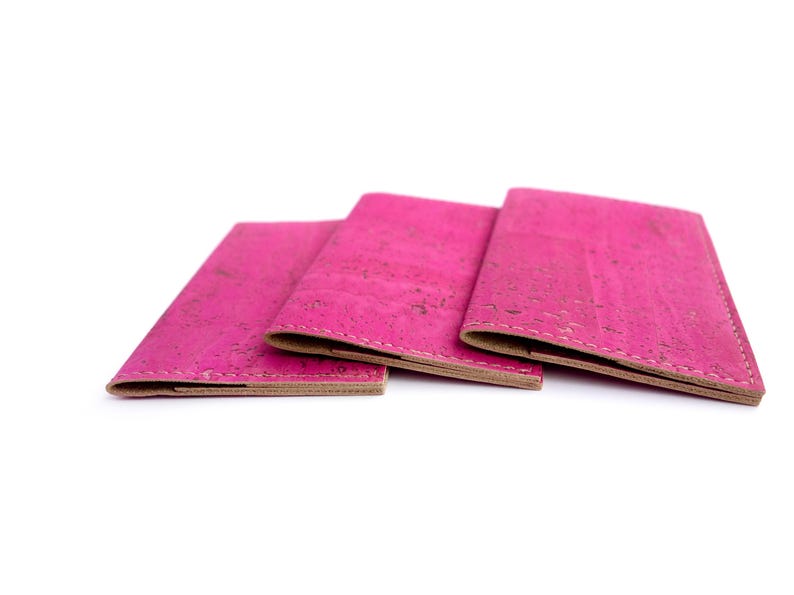 Pu&ograve; includere: Tre portafogli in sughero rosa acceso sono esposti su sfondo bianco. I portafogli sono aperti, rivelando un interno marrone chiaro. Il sughero ha un aspetto strutturato. I portafogli sono probabilmente per carte o contanti.