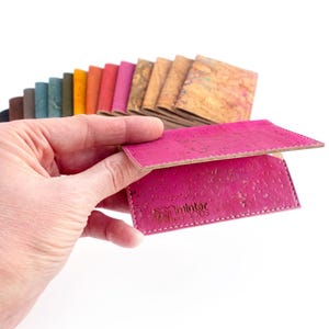 Pu&ograve; includere: Una mano tiene in mano un portafoglio in sughero rosa acceso, che mostra la sua superficie strutturata e le cuciture bianche. Sullo sfondo &egrave; visibile una selezione di campioni di sughero colorati. Il portafoglio ha il logo "minter woods".