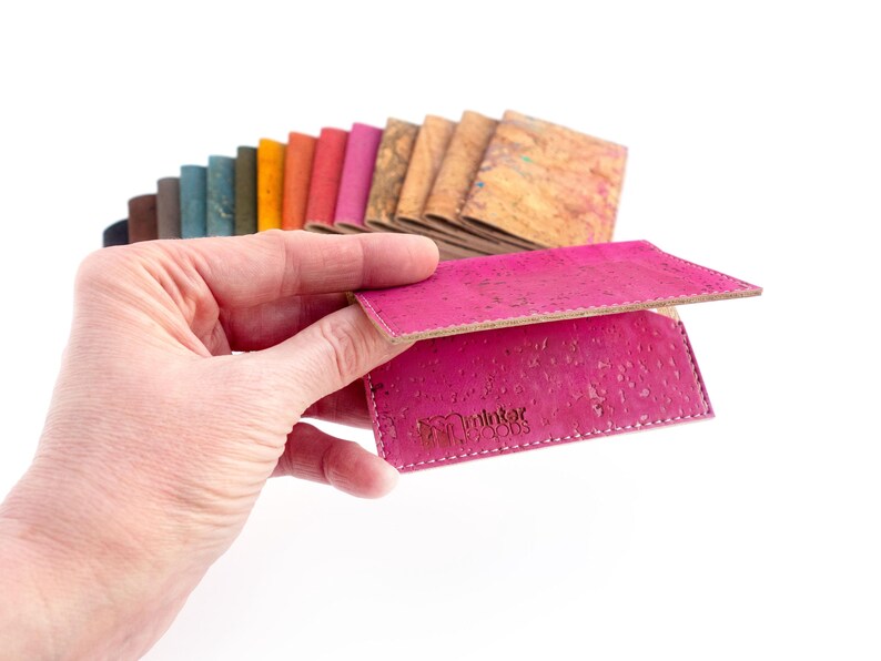 Pu&ograve; includere: Una mano tiene in mano un portafoglio in sughero rosa acceso, che mostra la sua superficie strutturata e le cuciture bianche. Sullo sfondo &egrave; visibile una selezione di campioni di sughero colorati. Il portafoglio ha il logo "minter woods".