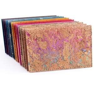 Pu&ograve; includere: Una pila di portafogli in sughero colorati. I portafogli sono in vari colori, tra cui marrone, blu, rosa e arancione. Il portafoglio anteriore ha un motivo unico con accenti viola e turchese. I portafogli sono impilati su uno sfondo bianco.