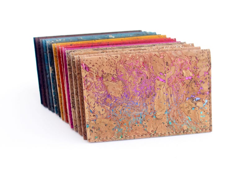 Pu&ograve; includere: Una pila di portafogli in sughero colorati. I portafogli sono in vari colori, tra cui marrone, blu, rosa e arancione. Il portafoglio anteriore ha un motivo unico con accenti viola e turchese. I portafogli sono impilati su uno sfondo bianco.