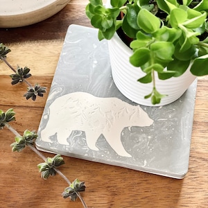 Op de afbeelding: Een grijze betonnen onderzetter met een wit beren silhouet ontwerp. De onderzetter ligt op een houten oppervlak naast een plant in een pot.