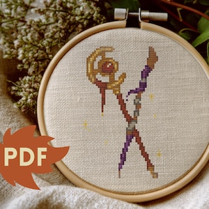 Puede incluir: Bordado de punto de cruz en un aro de madera. El dise&ntilde;o presenta una figura estilizada con un bast&oacute;n, en tonos marr&oacute;n, morado y dorado, con peque&ntilde;os acentos de estrellas amarillas. El texto "PDF" est&aacute; en un gr&aacute;fico en forma de hoja.