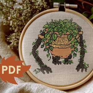 May include: Cross-stitch embroidery in a wooden hoop. The design features a terracotta pot with green foliage, supported by dark, vine-like arms and legs. The word "PDF" is on a leaf.