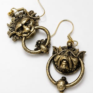 Puede incluir: Un par de pendientes de color oro antiguo. Cada pendiente presenta una cara detallada y esculpida con un elemento en forma de anillo. Los pendientes tienen un cierre de gancho y un dise&ntilde;o decorativo y texturizado. El fondo es blanco.