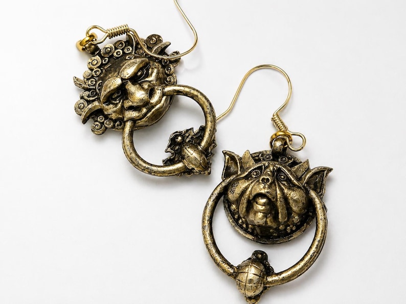 Puede incluir: Un par de pendientes de color oro antiguo. Cada pendiente presenta una cara detallada y esculpida con un elemento en forma de anillo. Los pendientes tienen un cierre de gancho y un dise&ntilde;o decorativo y texturizado. El fondo es blanco.