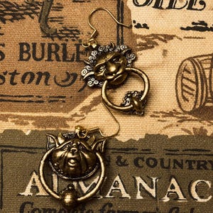 Puede incluir: Un par de pendientes dorados antiguos. Cada pendiente presenta un dise&ntilde;o detallado de rostro con un anillo debajo. Los pendientes se colocan sobre un fondo de estilo vintage con texto e ilustraciones.