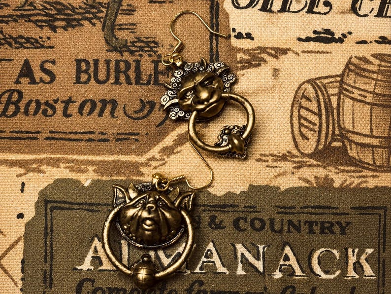 Puede incluir: Un par de pendientes dorados antiguos. Cada pendiente presenta un dise&ntilde;o detallado de rostro con un anillo debajo. Los pendientes se colocan sobre un fondo de estilo vintage con texto e ilustraciones.