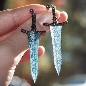 Peut inclure: Une paire de boucles d'oreilles uniques en forme de petites épées. Les lames sont bleu translucide avec un motif givré détaillé. Les gardes et les pommeaux sont marron foncé, et les boucles ont des tiges argentées.