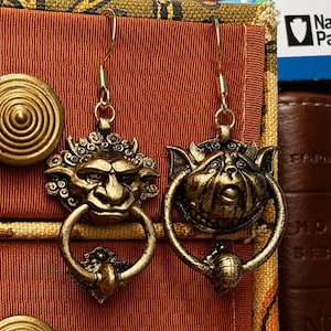 Puede incluir: Un par de pendientes antiguos de color dorado. Cada pendiente presenta una cara de duende detallada con un asa en forma de anillo. Los pendientes est&aacute;n suspendidos de un simple gancho dorado. El fondo incluye una tela roja y una pila de libros.