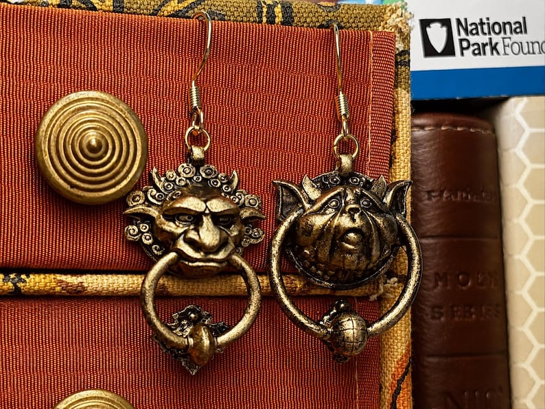 Puede incluir: Un par de pendientes antiguos de color dorado. Cada pendiente presenta una cara de duende detallada con un asa en forma de anillo. Los pendientes est&aacute;n suspendidos de un simple gancho dorado. El fondo incluye una tela roja y una pila de libros.