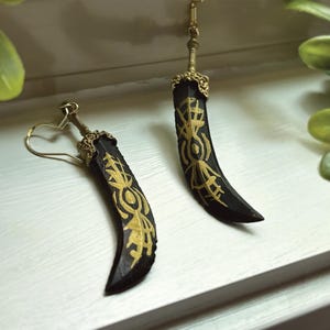 May include: A pair of black sword-shaped earrings with gold-colored filigree and decorative accents. The earrings have a curved blade design with intricate gold patterns. The ear hooks are gold-colored.