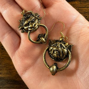 Puede incluir: Un par de pendientes de color oro antiguo. Cada pendiente presenta una cara estilizada y detallada con un elemento en forma de anillo. Los pendientes tienen un cierre de gancho. El dise&ntilde;o es intrincado y ornamentado, con una est&eacute;tica vintage.