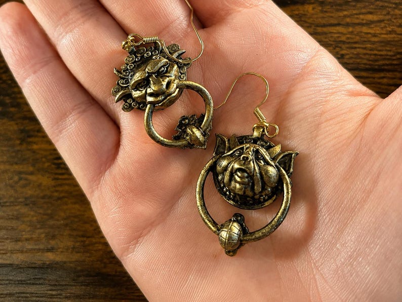 Puede incluir: Un par de pendientes de color oro antiguo. Cada pendiente presenta una cara estilizada y detallada con un elemento en forma de anillo. Los pendientes tienen un cierre de gancho. El dise&ntilde;o es intrincado y ornamentado, con una est&eacute;tica vintage.