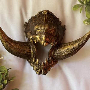 May include: A detailed, golden-colored mask with a fierce, animalistic design. The mask features prominent, curved horns and sharp teeth, suggesting a mythical creature. The overall aesthetic is ornate and textured, with a weathered appearance.