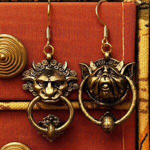 Puede incluir: Un par de pendientes dorados antiguos. Cada pendiente presenta una cara de duende detallada con un elemento en forma de anillo. Los pendientes est&aacute;n suspendidos de un gancho dorado. El fondo es una tela texturizada de color naranja rojizo.