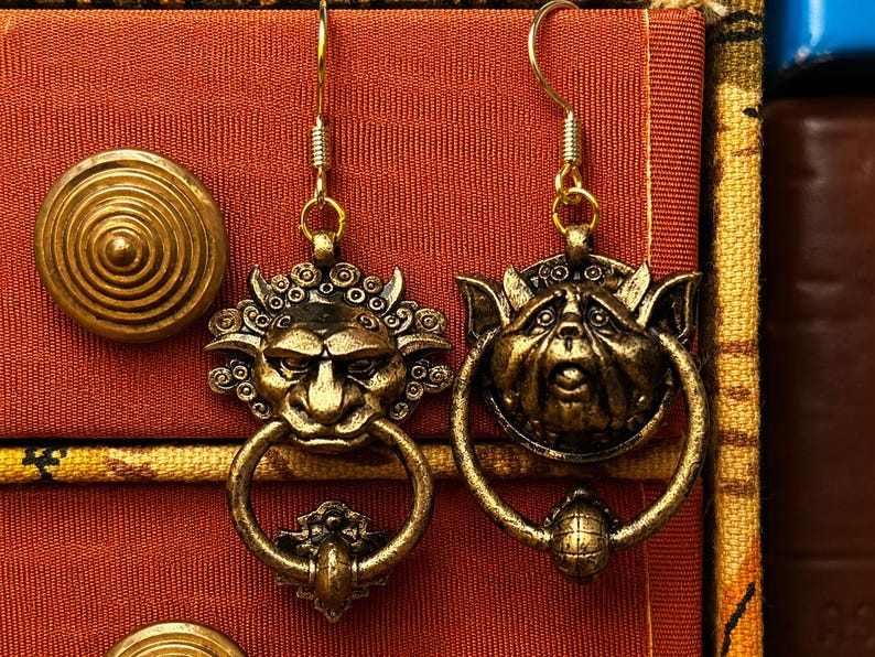Puede incluir: Un par de pendientes dorados antiguos. Cada pendiente presenta una cara de duende detallada con un elemento en forma de anillo. Los pendientes est&aacute;n suspendidos de un gancho dorado. El fondo es una tela texturizada de color naranja rojizo.