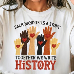 May include: White t-shirt with a graphic of six hands in various skin tones, each with a red heart. The text reads "EACH HAND TELLS A STORY" and "TOGETHER WE WRITE HISTORY" in brown and red.
