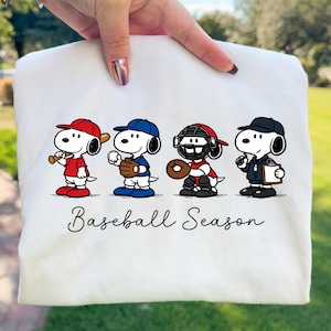 May include: White sweatshirt with a baseball-themed Snoopy design. Snoopy is depicted as a batter, pitcher, catcher, and coach. The phrase "Baseball Season" is written in cursive script beneath the cartoon characters.