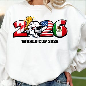 May include: White sweatshirt featuring a Snoopy character holding a trophy and soccer ball. The number 2 is red with a Canadian flag, 0 is a soccer ball, 2 is an American flag, and 6 is a Mexican flag. The text "WORLD CUP 2026" is below the numbers.
