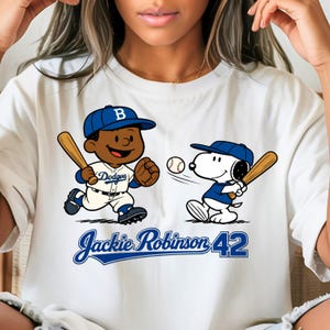 May include: White t-shirt featuring a cartoon illustration of a baseball player and Snoopy playing baseball. The baseball player is wearing a Dodgers uniform and a blue cap. Snoopy is also wearing a blue cap and uniform. The text "Jackie Robinson 42" is below the illustration.